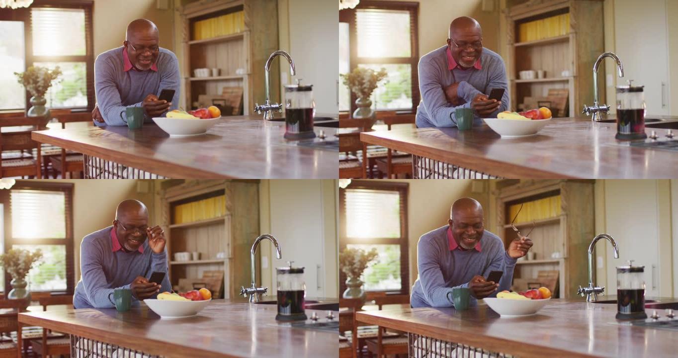 非裔美国人高级男子使用智能手机靠在厨房柜台上，摘下眼镜，微笑着