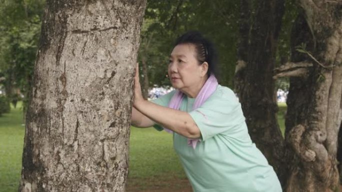 POV手持中枪: 超重的亚洲高级女性在公园跑步后伸展手臂并推树进行锻炼。老运动女人扭动身体，早上感到