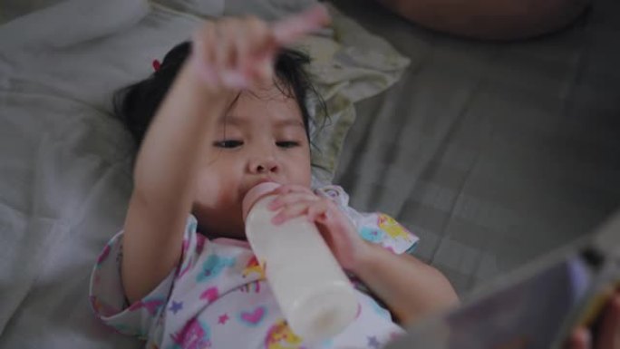 亚洲女婴一边喝着牛奶，一边和祖母在卧室看智能手机