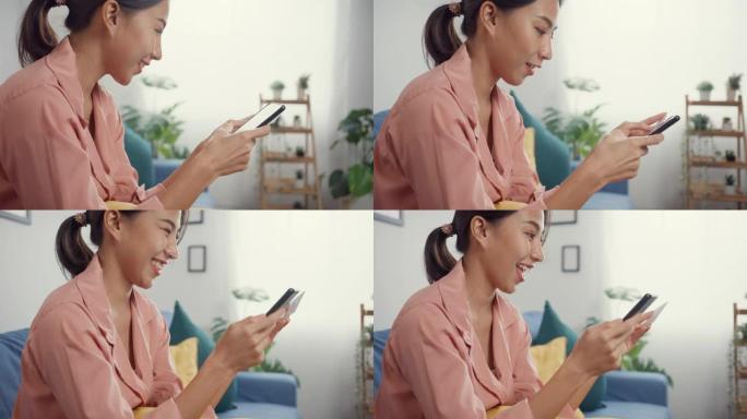 快乐的年轻亚洲女士使用手机订购在线购物产品，用信用卡支付账单，坐在家里客厅的沙发上。
