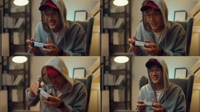 快乐的亚洲年轻人穿着灰色连帽衫，保持操纵杆控制器兴奋和专注，同时晚上坐在家里客厅的沙发上玩电子游戏。
