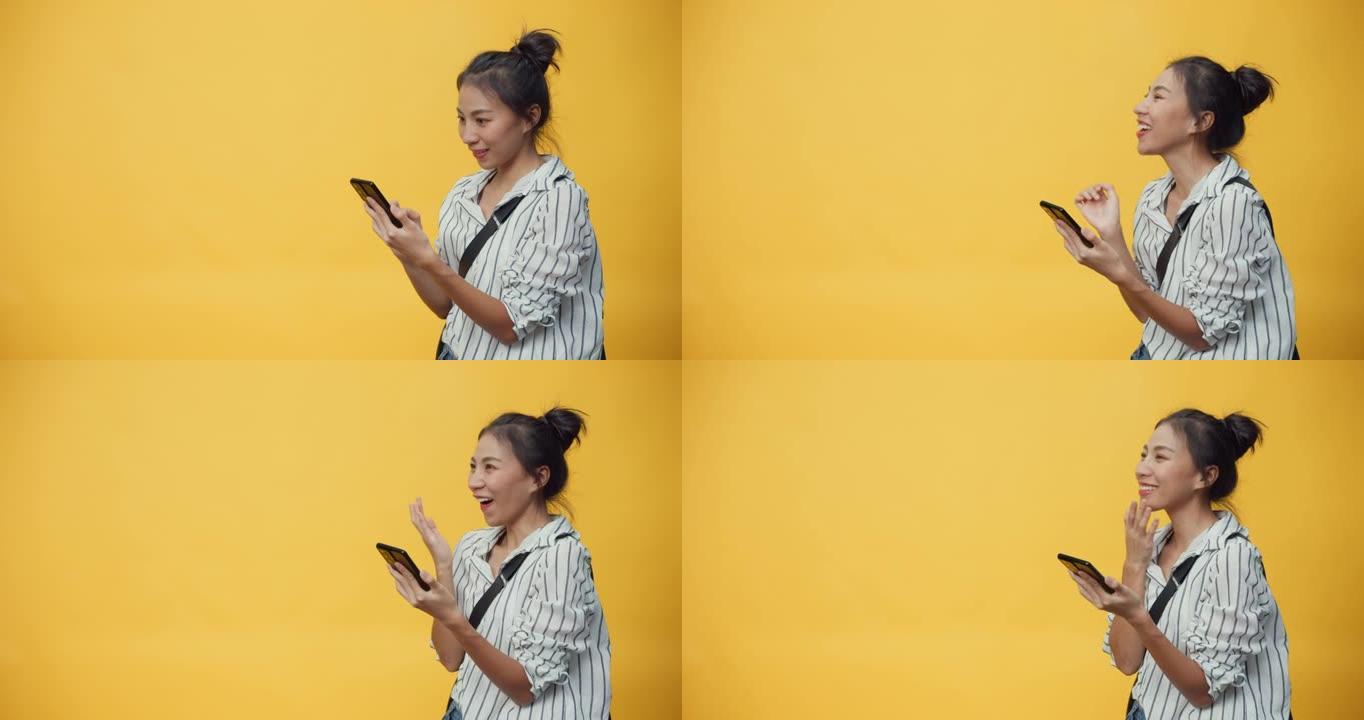 亚洲青年休闲女孩微笑享受打字智能手机站在孤立的黄色空白空间背景与微笑给予标志呈现惊喜退出成功购买在线