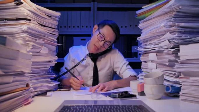 32岁的亚洲商人戴着眼镜忙于文书工作，由于工作到很晚的晚上在办公室工作过多而对压力感到不满。工作到很