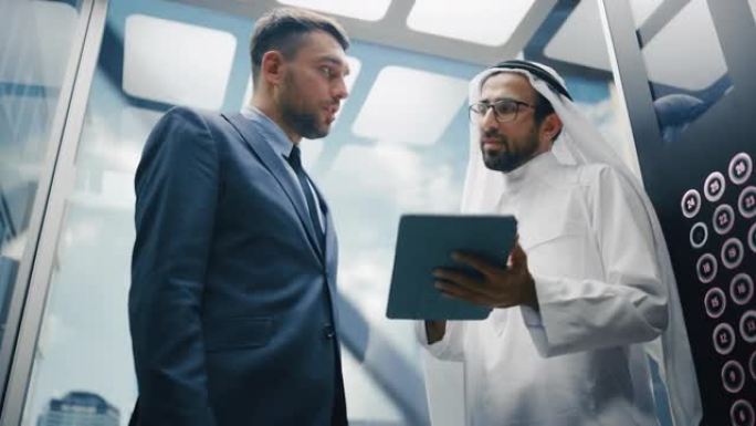 欧洲商人在现代商务中心乘坐玻璃电梯到办公室时与阿拉伯投资伙伴交谈。国际公司员工在电梯中使用平板电脑。
