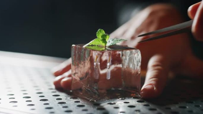调酒师的特写镜头是将薄荷和镊子放在专业的水晶冰块中，为酒吧或迪斯科俱乐部的顾客准备酒精鸡尾酒。