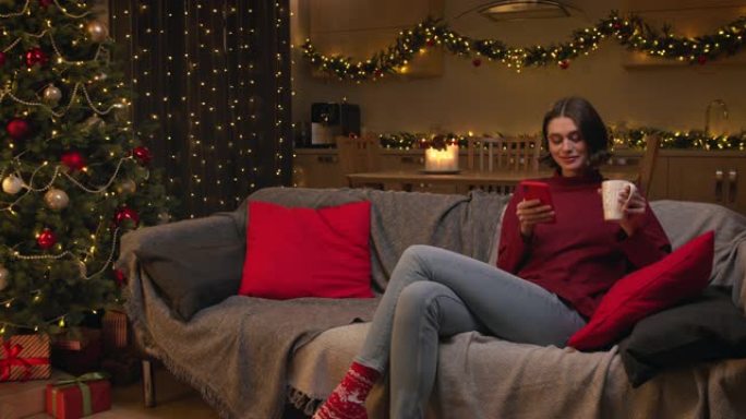 一位迷人的年轻女子坐在沙发上，用她的红色智能手机在网上购物，喝杯茶，她在一个有圣诞灯和装饰圣诞树的房