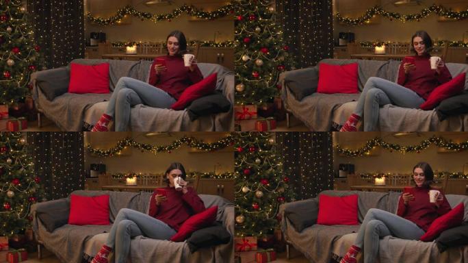 一位迷人的年轻女子坐在沙发上，用她的红色智能手机在网上购物，喝杯茶，她在一个有圣诞灯和装饰圣诞树的房