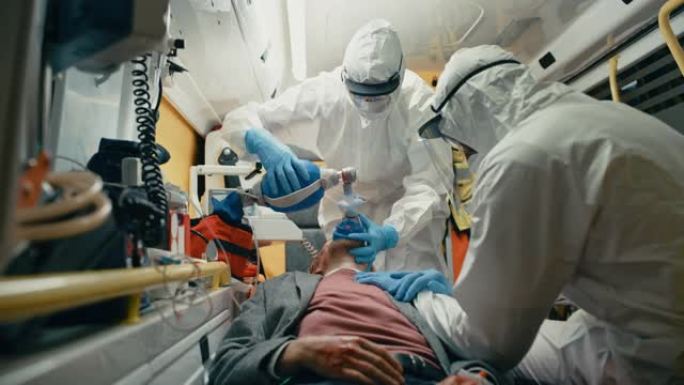 身穿一次性工作服的急救护理人员在送往医院的途中为受伤的病人提供医疗帮助。急救助理在救护车上戴上无创通