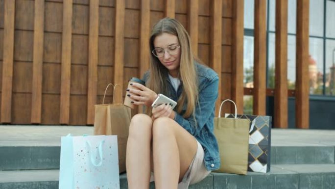 年轻女子使用智能手机坐在户外，带着购物袋在夏日放松