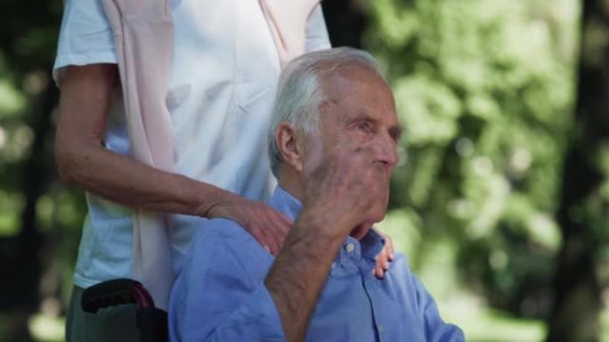 成熟妻子的电影特写镜头正在拥抱，这是永远永恒的爱让轮椅上的丈夫残疾的标志，在绿色公园的镜头里微笑着。
