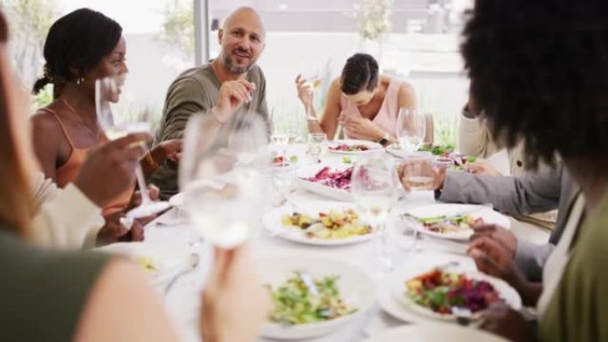 多元化的团体在享受美食和美酒的聚会上大笑。人们吃饭和喝酒，在一张大餐桌上一起社交。朋友在家里的餐厅吃