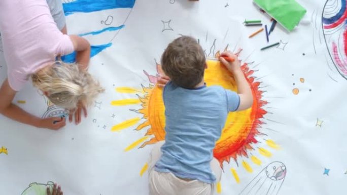 从上面看，幼儿和朋友在幼儿园的地板上在室内绘制彩色太空图片。孩子们正在创造性地画科幻画，在一张大张纸