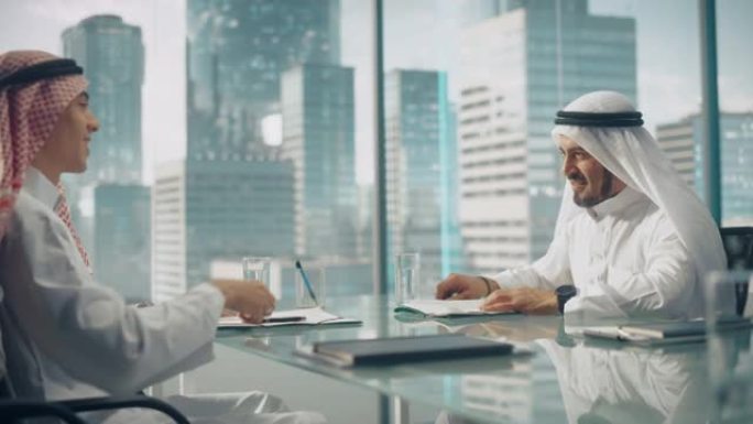 两个快乐的阿联酋商人穿着白色传统的坎杜拉坐在办公室并签署合同。商业伙伴就有利可图的投资握手。沙特、阿