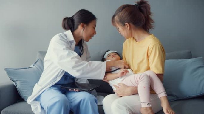 年轻的亚洲女儿科医生举行听诊器检查小女孩病人去看医生，母亲坐在家里客厅的沙发上。