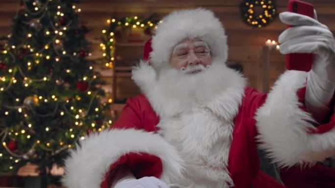 圣诞老人在他装饰精美的小屋里，圣诞树在他身后，正在他的红色智能手机上与某人视频聊天