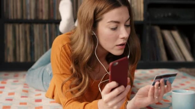年轻的白人妇女用手机拿着信用卡放松在家听音乐