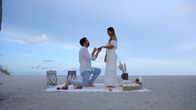 在海滩上浪漫野餐时，有爱心的男人跪在地上，问惊讶的女人是否愿意嫁给他