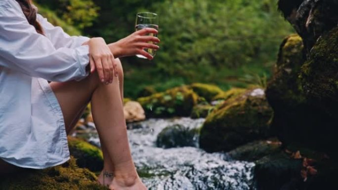 SLO MO女人坐在湍急的溪流旁的岩石上时，拿着一杯纯净的矿泉水