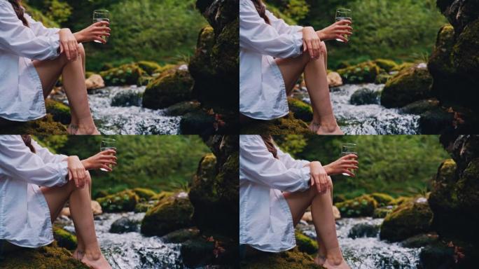 SLO MO女人坐在湍急的溪流旁的岩石上时，拿着一杯纯净的矿泉水