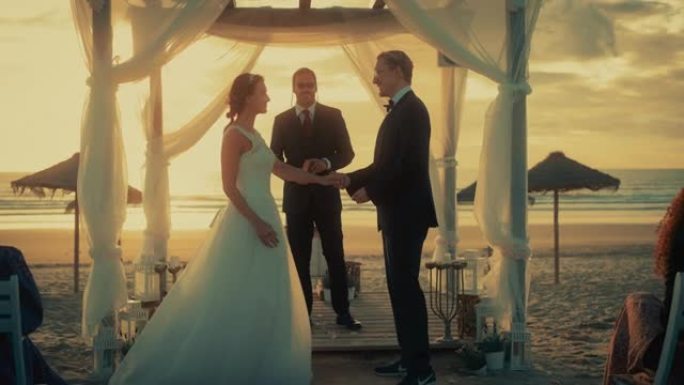 美丽的新娘和新郎在日落时在海洋海滩举行的户外婚礼上。一对夫妇结婚、交换戒指、亲吻和与多民族朋友分享庆