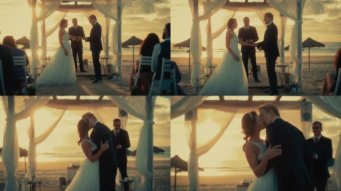美丽的新娘和新郎在日落时在海洋海滩举行的户外婚礼上。一对夫妇结婚、交换戒指、亲吻和与多民族朋友分享庆