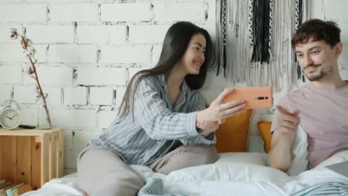 亚洲妇女使用智能手机微笑显示屏幕给丈夫在家里床上使用笔记本电脑