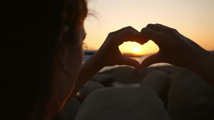 一位年轻快乐微笑的黑发女人游客的真实特写镜头正在用手做心形，同时在海边享受暑假假期，日落时分有海上游