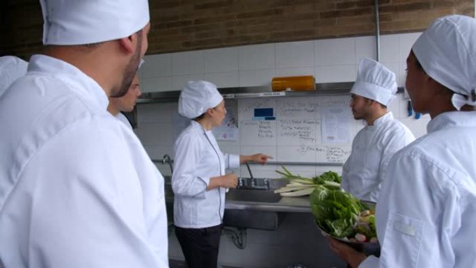 首席女厨师指着墙上计划的菜单，并在餐厅的厨房为她的团队分配任务