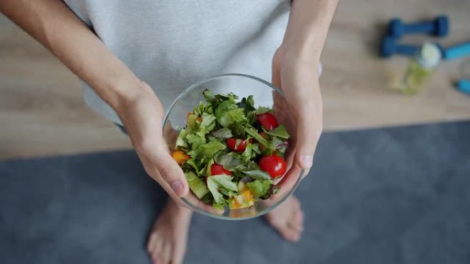 男人的手站在家里的瑜伽垫上拿着一碗蔬菜沙拉的俯视图