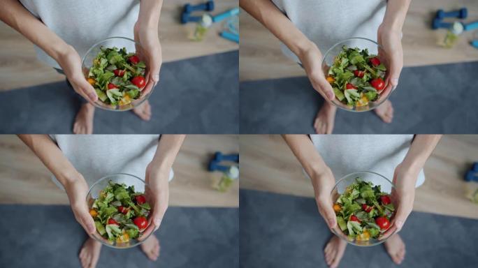 男人的手站在家里的瑜伽垫上拿着一碗蔬菜沙拉的俯视图