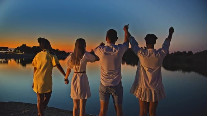 SLO MO四个朋友在黄昏时在湖边的海滩上举起手来