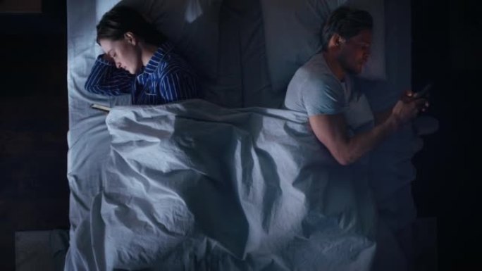 顶视图公寓卧室: 年轻夫妇躺在床上，都使用智能手机。两口之家使用手机浏览社交媒体，交流，搜索互联网，