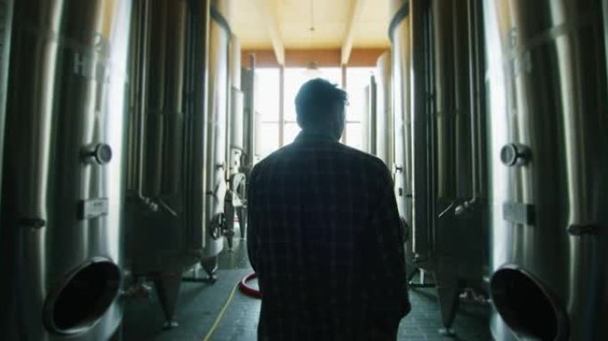 一个快乐的成功的农民或酿酒师正走在酒罐中间，并在现代化的酿酒设施或酿酒厂中检查高质量葡萄酒生产的发酵