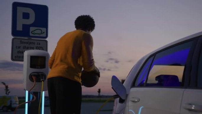SLO MO Young篮球运动员在黄昏时使用智能手机在停车场为电动汽车充电