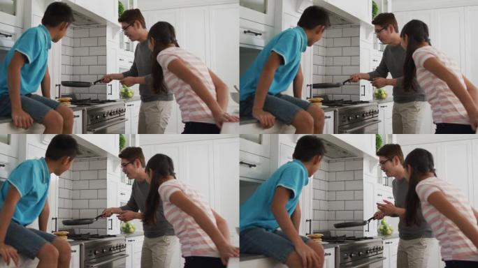 快乐的亚洲父亲带着儿子和女儿在厨房里，一边看一边在煎锅里翻煎饼
