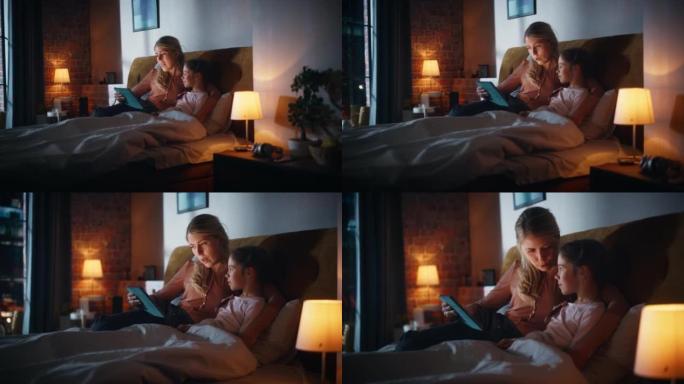 母亲在睡觉前从平板电脑上给她可爱的女儿读童话。年轻的妈妈照顾小女孩，在家阅读在线故事。手持镜头。
