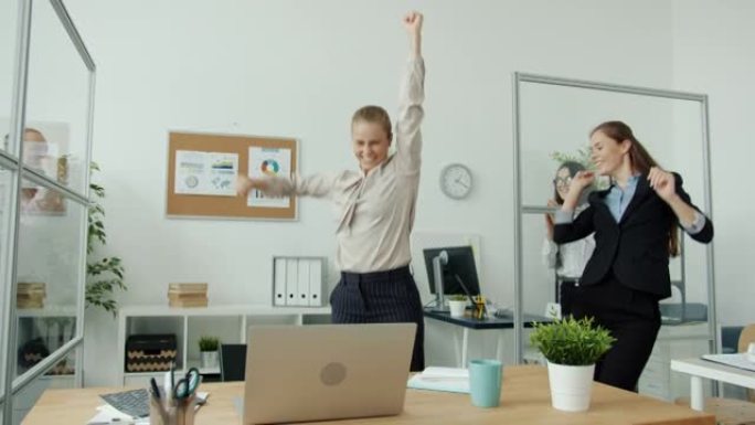 兴奋的女商人使用笔记本电脑，然后得到好消息，并在办公室与同事跳舞