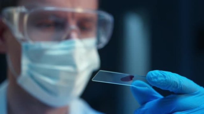 在现代实验室中，戴着防护面罩和眼镜的科学家的特写镜头看着带有血滴的显微镜载玻片