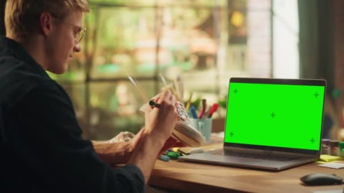 男性设计师在带有绿屏模拟色度键的笔记本电脑前的鞋子上绘画。十几岁的自由职业者在他的工作室设计独特的鞋