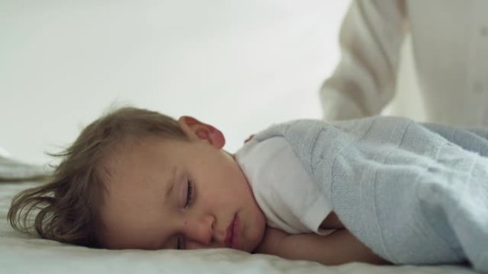 母亲的真实特写是用柔软的毯子盖住她蹒跚学步的男婴，同时白天在托儿所的婴儿床里睡觉。家庭，舒适，关怀，