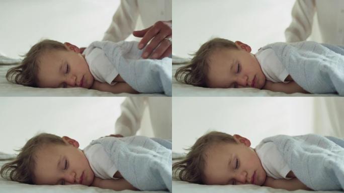母亲的真实特写是用柔软的毯子盖住她蹒跚学步的男婴，同时白天在托儿所的婴儿床里睡觉。家庭，舒适，关怀，