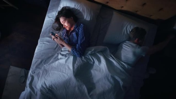 顶视图卧室公寓: 年轻女子晚上在床上使用智能手机，当时她的女性伴侣试图在旁边入睡。夫妻吵架，争论。社