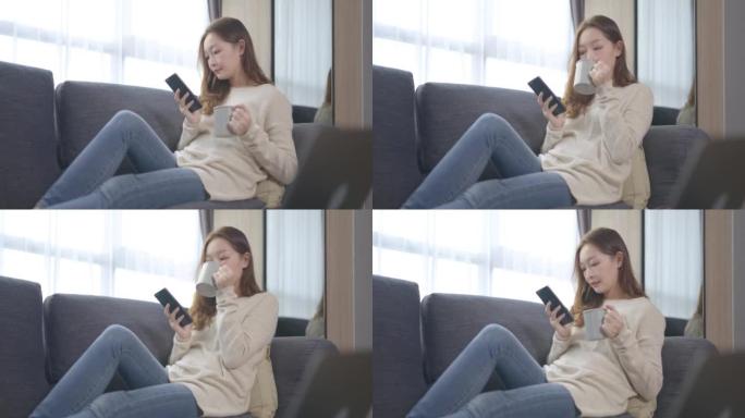 美丽的亚洲年轻女子年龄36岁，通过手机工作和网上购物，同时享受一杯咖啡，坐在沙发上放松，早上在家。成