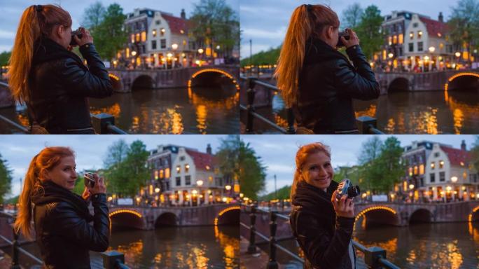 在阿姆斯特丹度假的女性游客，晚上用老式模拟相机拍摄运河上的照明桥梁，微笑着对着相机