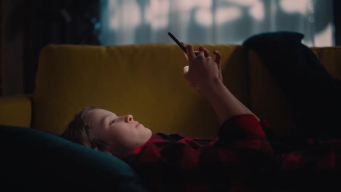 可爱的小男孩躺在黑暗舒适的房间的沙发上，在家使用智能手机。快乐的青少年在线浏览内容，在社交媒体上与朋