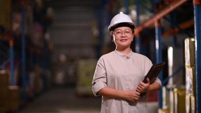 亚洲华人管理成熟的女人看着相机微笑着在仓库里戴着白色安全帽拿着数字平板电脑