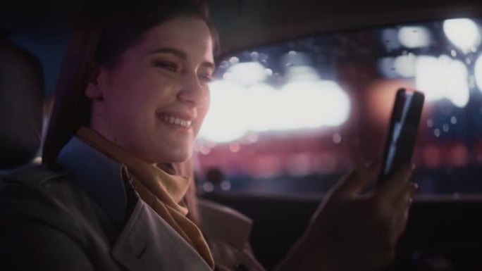 快乐的女性晚上坐出租车后座回家。美丽的女人乘客在带有工作霓虹灯的城市街道上的转运车上通过智能手机进行