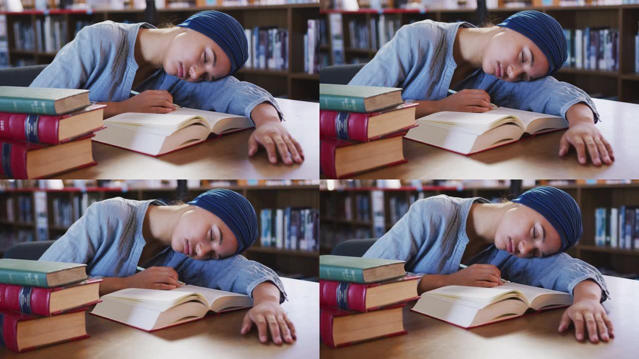 穿着蓝色头巾的亚洲女学生坐在打开书本的书桌前睡觉