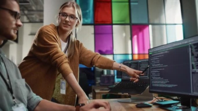 男性程序员在台式计算机上向时尚办公空间中的女性首席开发人员展示代码。高加索女人用有用的编码技巧帮助她