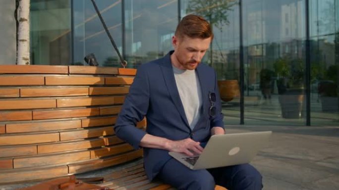 一位穿着蓝色西装的年轻有魅力的商人正坐在商务中心附近的长凳上，在笔记本电脑上打字